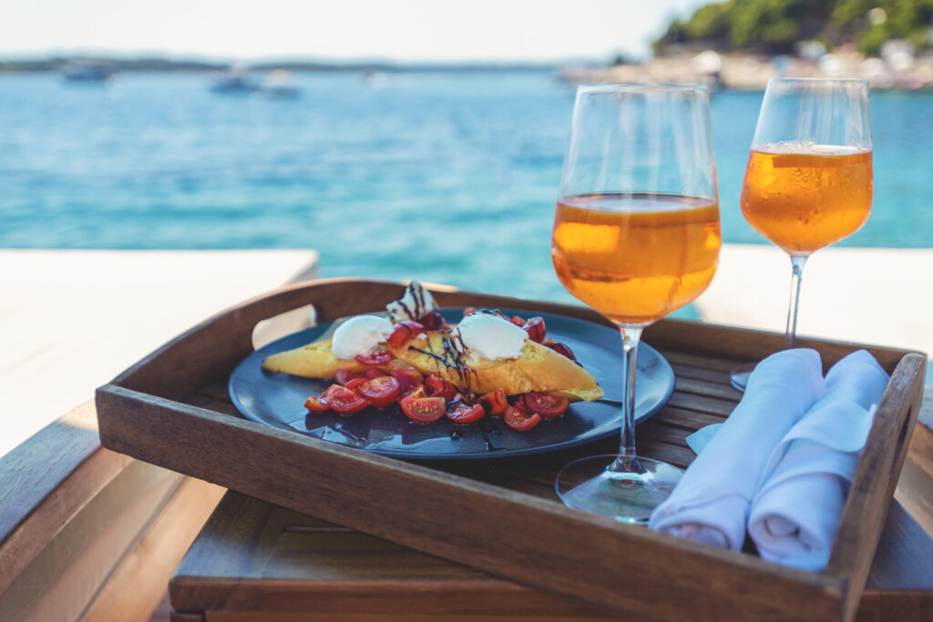 Two deck chairs with a Spritz cocktail and a meal of bruschetta and burrata on a deck over the ocean. Turquoise ocean is crystal clear.